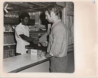 Printed black and white photograph of USS Intrepid's ship store with a sailor buying a carton o…