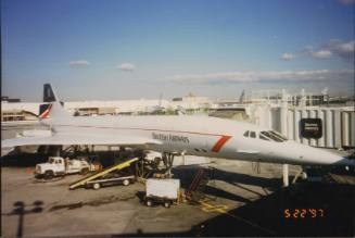 Color image of the exterior of a British Airways Concorde parked at an aiport gate with extende…