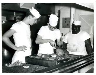 Black and white photograph of one cook carving a roast beef and two cooks looking on