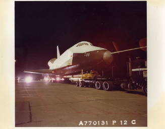 Color photograph of Enterprise on the back of a truck waiting on an airport taxi-way