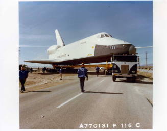 Color photograph of Enterprise being towed around a corner of of a street with people walking a…