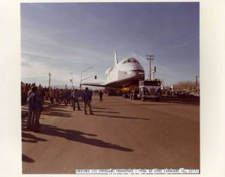 Color photograph of Enterprise being towed by a truck with people observing on the side of the …