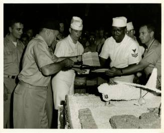 Black and white photograph of the ship's captain cutting a large cake and passing out pieces