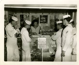 Black and white photograph of enlisted men standing at a USO counter with a woman behind the de…