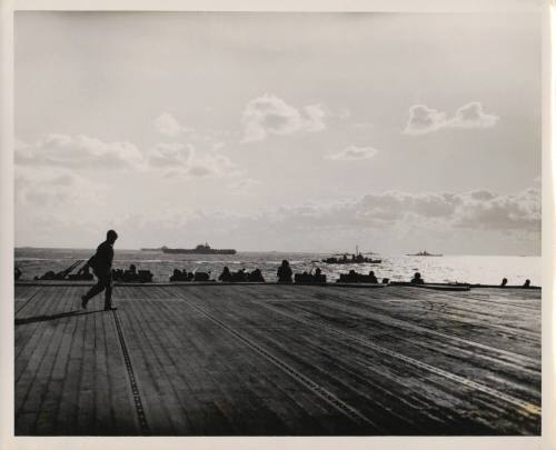 Black and white photograph of a man walking across USS Intrepid's flight deck, with other ships…