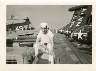 Black and white photograph of sailor posing on a flight deck tractor, with aircraft in the back…