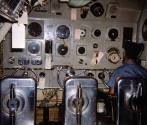 Printed color photograph of USS Growler's control room with a sailor seated at a panel of equip…