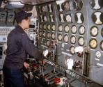 Color photograph of a crew member standing in front of the engine room control booth on the sub…