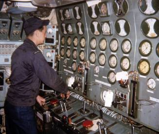 Color photograph of a crew member standing in front of the engine room control booth on the sub…