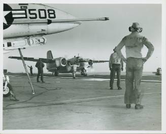 Black and white photograph of an air taxiing down the flight deck with flight deck personnel st…