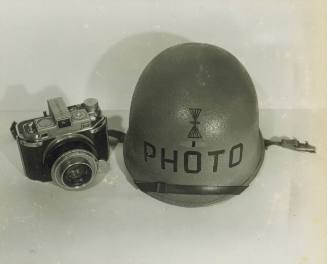 Black and white photograph of a camera and hard helmet with a photographer’s mate rating on it