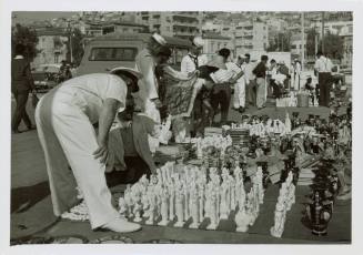 Black and white photograph of officers and enlisted men looking at souvenirs along a street in …