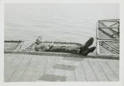 Black and white photograph of an enlisted man sleeping in a safety net off the side of the flig…