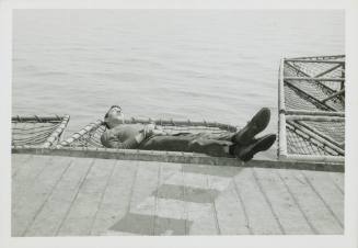 Black and white photograph of an enlisted man sleeping in a safety net off the side of the flig…