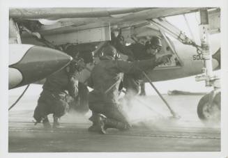 Black and white photograph of three crew members attaching an aircraft bridle to a plane on the…