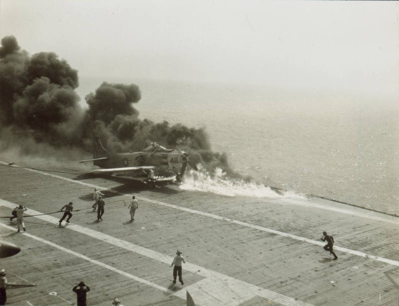 Black and white photograph of an AD-6 Skyraider engulfed in flames with crew members running to…