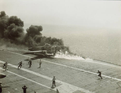 Black and white photograph of an AD-6 Skyraider engulfed in flames with crew members running to…