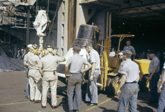 Color photograph of the practice space capsule for the Mercury 7 recovery surrounded by men on …