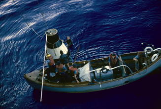 Color photograph of a motor boat in the water beside a practice Mercury space capsule