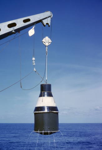 Color photograph of practice space capsule being lifted out of the ocean with a crane