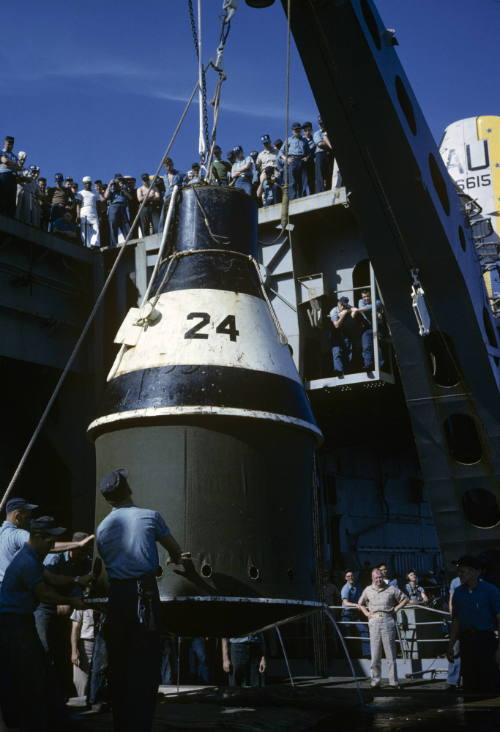 Color photograph of practice space capsule on starboard aircraft elevator with men watching fro…