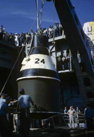 Color photograph of practice space capsule on starboard aircraft elevator with men watching fro…