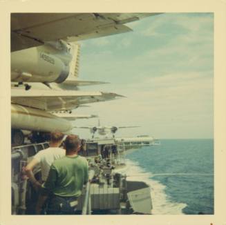 Color photograph of two crew standing on the catwalk with aircraft on the flight deck above