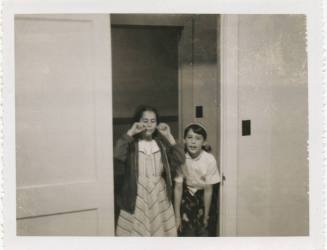 Black and white image of two girls standing in doorway, the girl on the left has her hands rais…