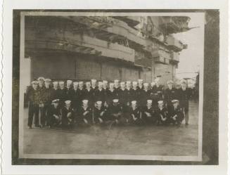 Black and white image of a Gunnery Department division, enlisted men stand and kneel in a line …