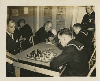 Black and white image of four men sitting around a table with two chess sets at the center of t…