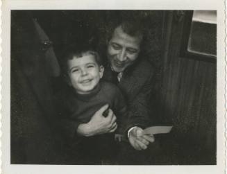 Black and white image of a man sitting down and holding a child with one hand and a letter in a…