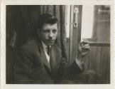 Black and white image of a man in a suit smoking a cigarette while seated, interior door to a t…