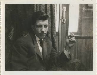 Black and white image of a man in a suit smoking a cigarette while seated, interior door to a t…