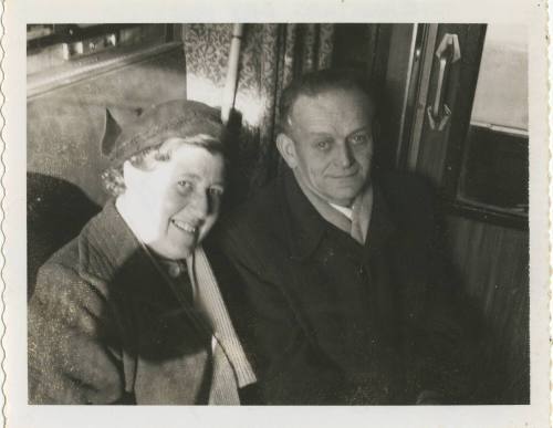 Black and white image of a man and woman sitting on a train wearing coats, interior train door …