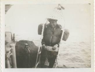 Black and white image of an officer in a life jacket sitting in a highline chair, package on la…