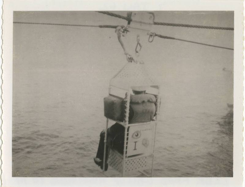 Black and white image of a man seated in a highline chair wearing a life jacket over water