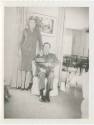 Black and white image of a man seated in a chair and a woman standing next to him, painting han…