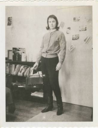 Black and white image of a woman standing in front of a wall with cards on it, a book shelf is …