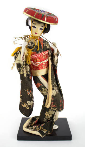Front view of doll depicting a Japanese woman wearing black floral kimono, red belt and hat sta…