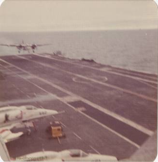 Color photograph of a plane landing on the aircraft carrier USS Intrepid's flight deck, with ot…