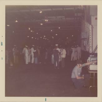 Color photograph showing crew members and civilians talking in groups on the hangar deck of USS…