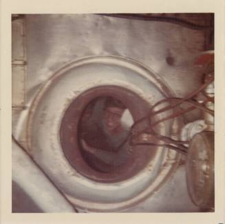 Color photograph of a round opening in the side of a ship's boiler, with a smiling man inside t…