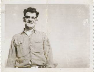 Black and white image of Jack Standish smiling at the camera, he has dark hair and wears glasse…