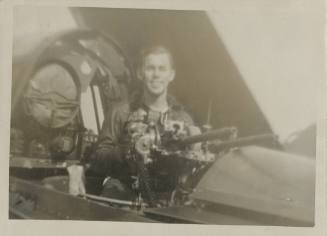 Black and white photograph of a man standing in open gunner's compartment of a Curtiss SB2C Hel…