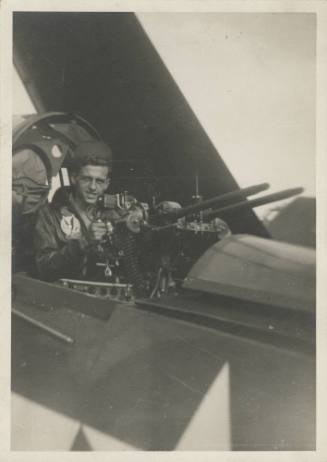 Black and white photograph of a man stansing in an open gunner's compartment of a Curtiss SB2C …