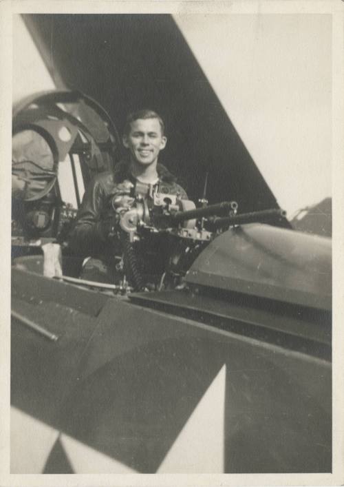 Black and white photograph of a man standing behind the guns in an open cockpit of a Curtiss SB…