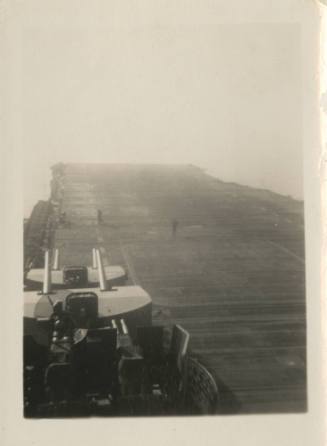 Black and white image looking at the aft half of the flight deck, anti-aircraft guns are visibl…