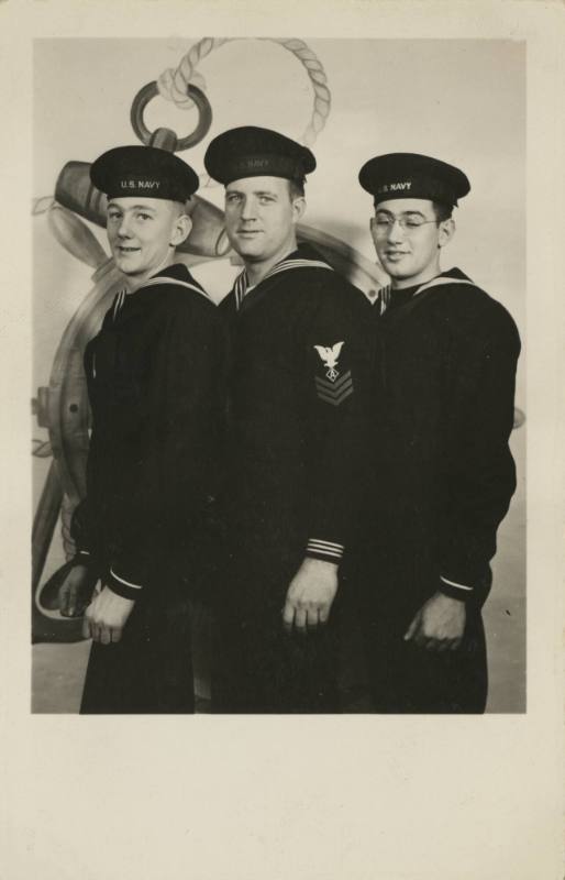 Black and white image of three men in US Navy dress blue uniforms standing in front of graphic …