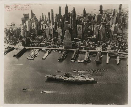 Black and white photograph of USS Intrepid sailing down the East River with small boats on eith…