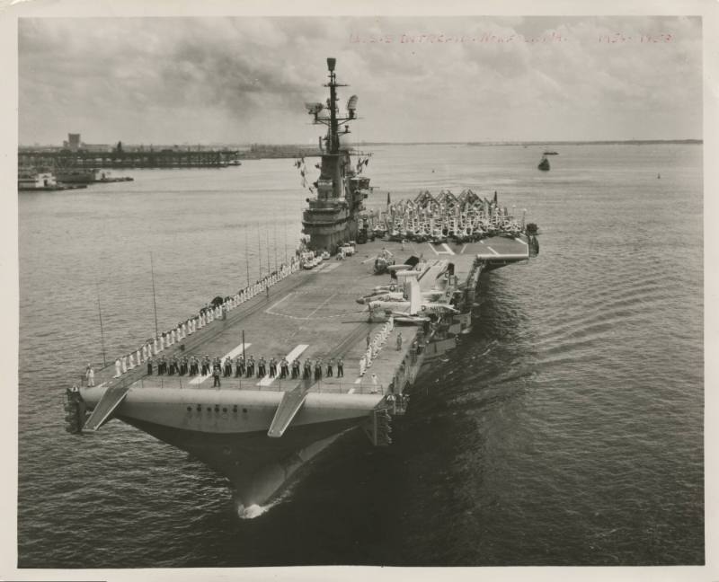 Black and white photograph of aircraft carrier USS Intrepid sailing with crew standing along po…